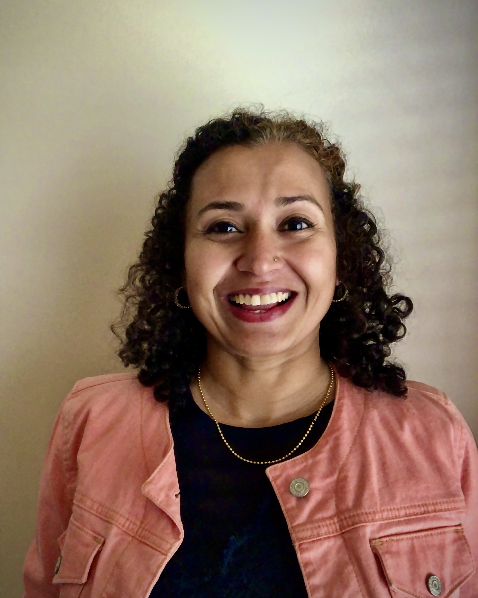 Chandni Krishnan laughing, in a coral jacket and a gold necklace, against a pale wall